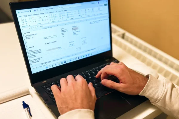 En bærbar computer med et regnskabsprogram åbent og to hænder, der skrive på computerens keyboard. Der er også en notesbog og kuglepen.