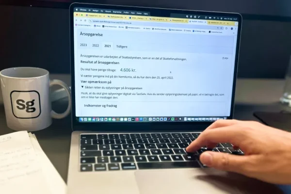 En laptop, der viser resultatet af en årsopgørelse. Der er også en notesbog, en Skatteguiden kop og en hånd.