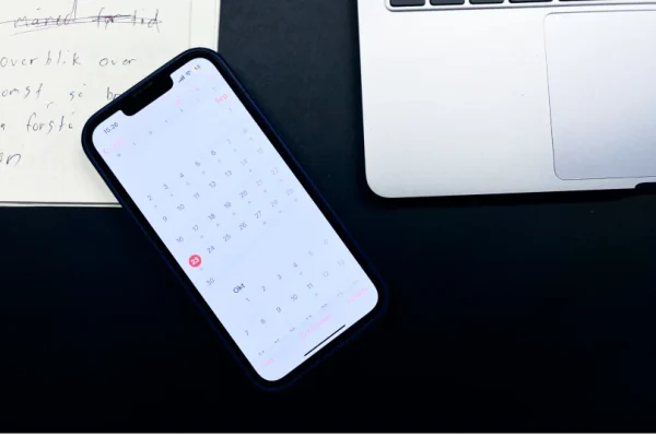En iPhone med kalender-appen åbent, ved siden af en laptop og en notesbog.