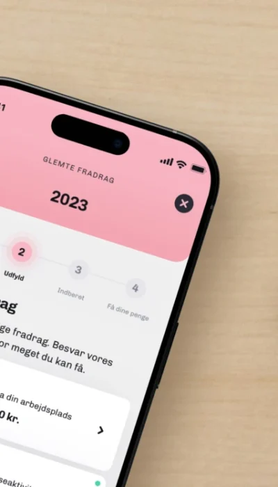 En iPhone på et hvidt træbord, der viser funktionen "Glemte Fradrag" i Skatteguidens app ved siden af en notesbog og en kuglepen.