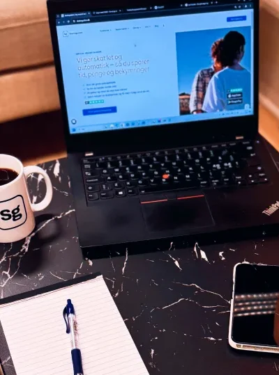 Bord med bærbar, der visser Skatteguidens hjemmeside, ved siden af en kaffekop, notesbog og en plante.