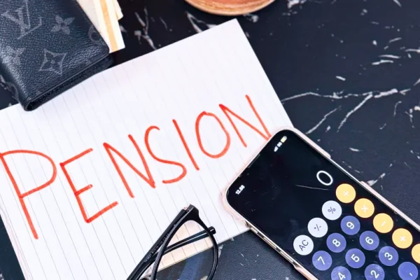 Der står "Pension" på et stykke papir, der ligger på et bord ved siden af en mobil med lommeregner, et par briller og en pung med kontanter i.