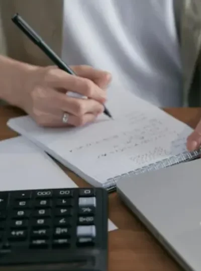 Billede af en person, der laver beregninger i en notesbog, der ligger bag ved en notesbog og en bærbar.