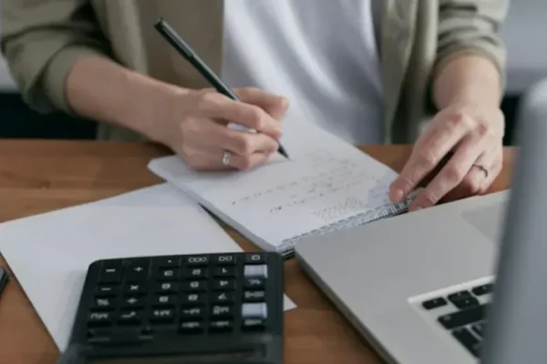 Billede af en person, der laver beregninger i en notesbog, der ligger bag ved en notesbog og en bærbar.