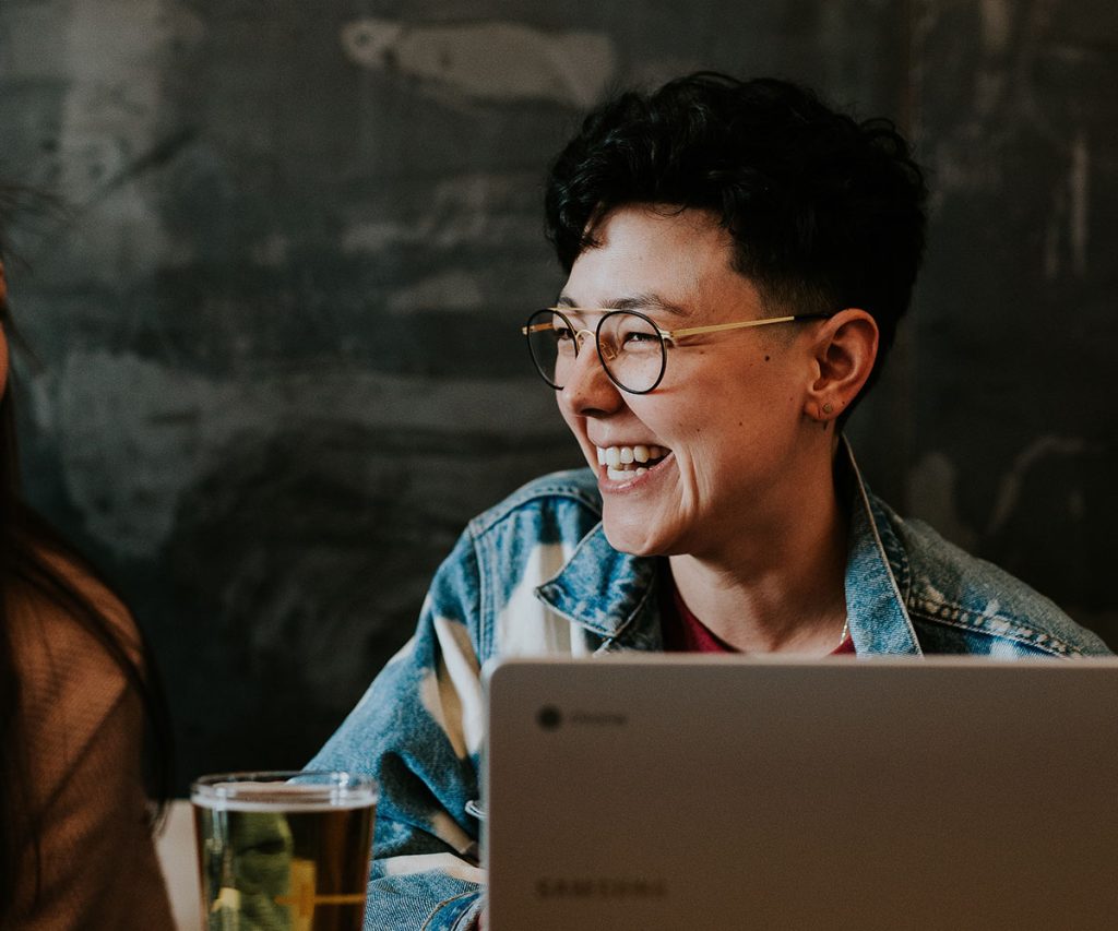 Smilende person med briller, der er engageret i en samtale om et bord. Har sin laptop åben.